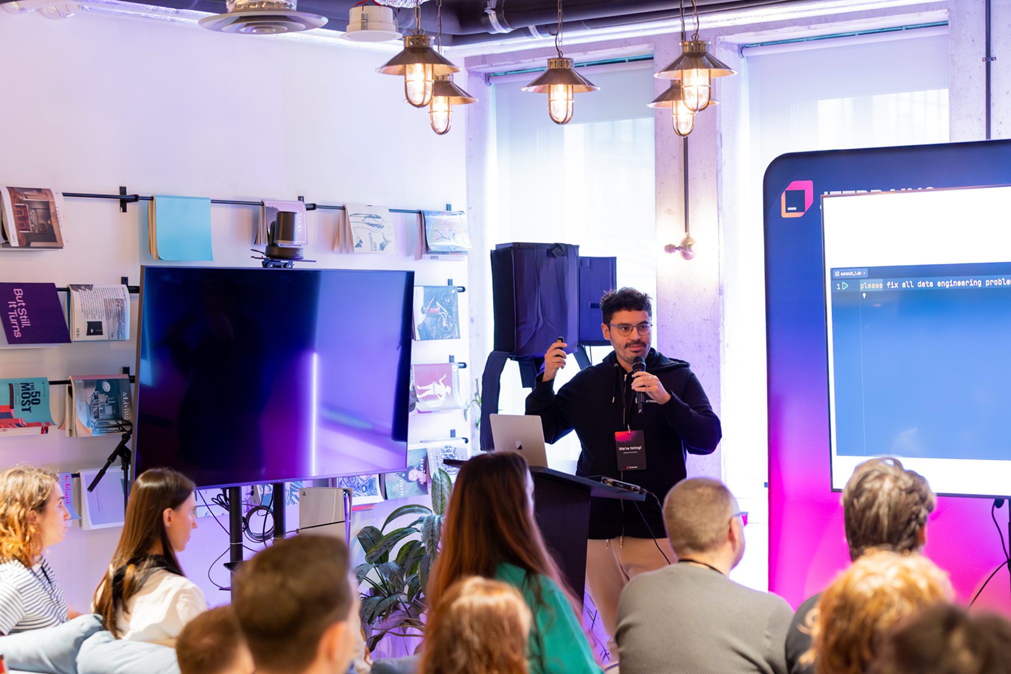 A speaker presenting with a microphone to a seated audience at a JetBrains meetup, with a large code screen behind them.